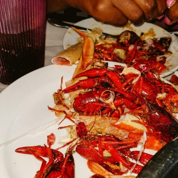 A white plate piled high with red crawfish shells and discarded pieces, with a drink and hands in the background.