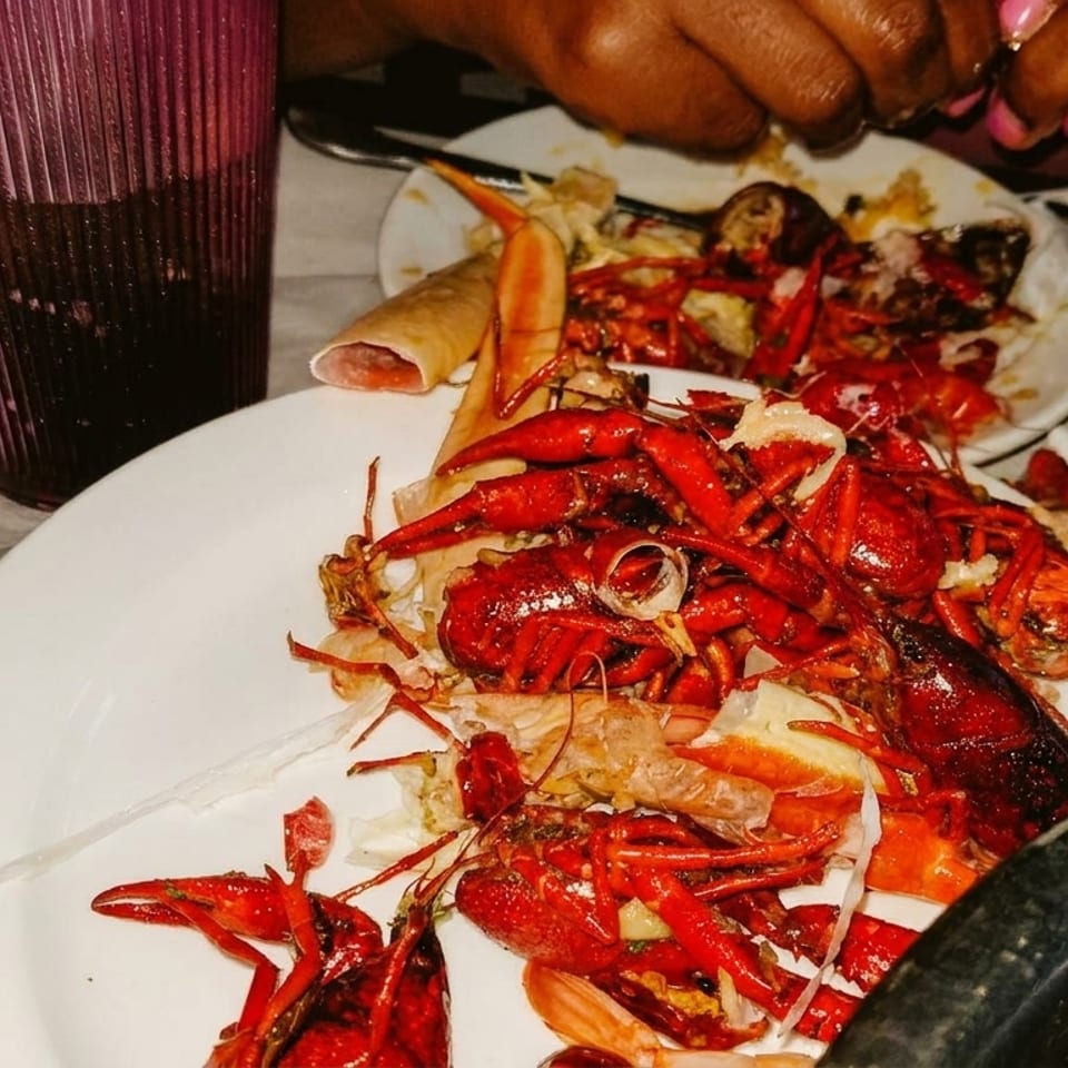 A white plate piled high with red crawfish shells and discarded pieces, with a drink and hands in the background.